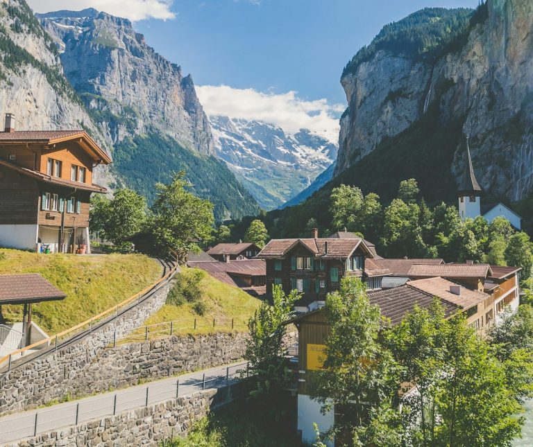 Kontakt Daten Berglandschaft mit einem Dorf, umgeben von hohen Felsen und schneebedeckten Gipfeln.